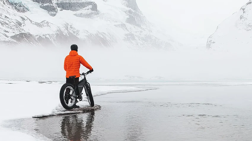 man in orange jacket standing next to sondors electric bicycle surrounded by a snowy landscape